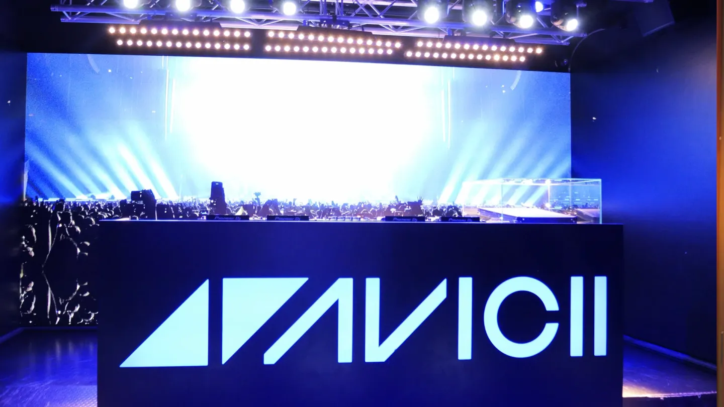 A stage setup at the Avicii Experience museum featuring a large illuminated AVICII logo on the front of a DJ booth, with bright blue lights and a backdrop showing a concert crowd under vivid spotlights.